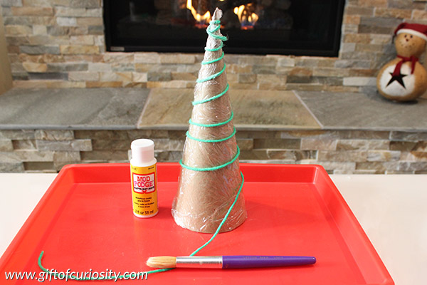 Build this cute Christmas tree craft for the holidays using green yarn and Mod Podge. Then trip your tree with mini lights, jeweled stickers, and more! || Gift of Curiosity